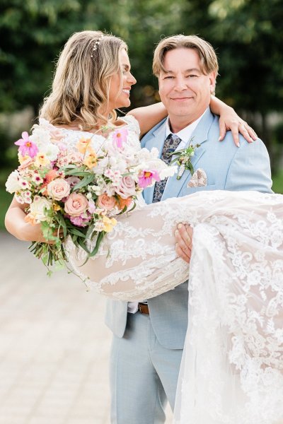 Groom picks up bride standing in park trees in background