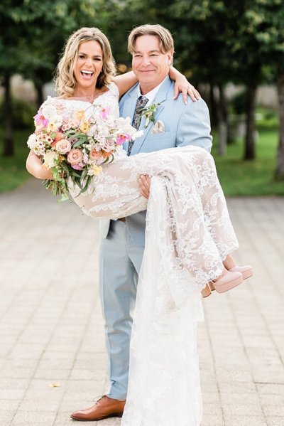 Groom picks up bride standing in park trees in background