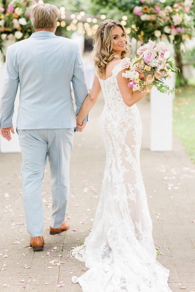 Bride from behind she looks over shoulder and holds grooms hand holding hands in park forest setting