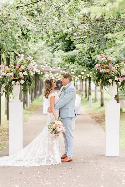 Bride and groom either side of flower pillar in forest park setting trees in background they hug