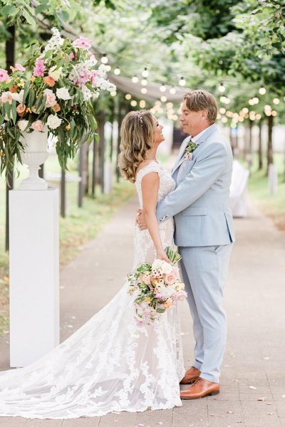 Bride and groom either side of flower pillar in forest park setting trees in background they hug