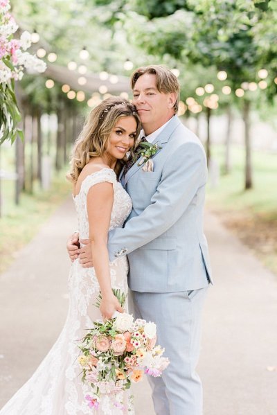 Bride and groom either side of flower pillar in forest park setting trees in background and lights they hug