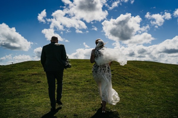 Bride and groom walk in the sun on the grass blue sky clouds walking away from camera veil blowing in the wind