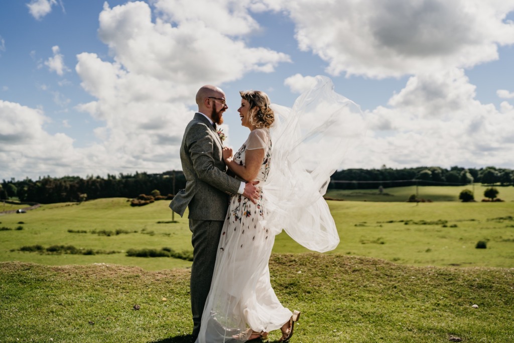 Bride and groom standing on grass skyline in background holding each other