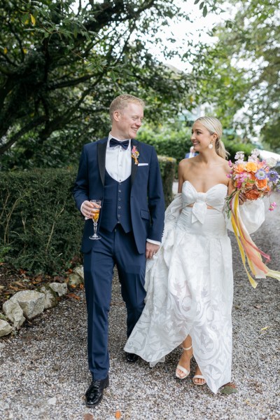 Bride and groom walk along pathway groom holds glass of champagne and bride holds bouquet