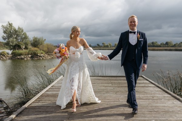 Bride and groom walk along the boardwalk lake in background they hold hands