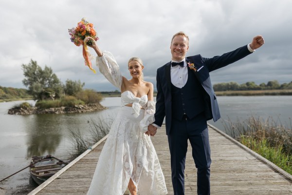 Bouquet in the air holding hands with groom bride smiles lake in background