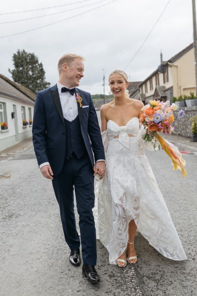 Bride and groom walk down road orange bouquet flowers they smile and laugh