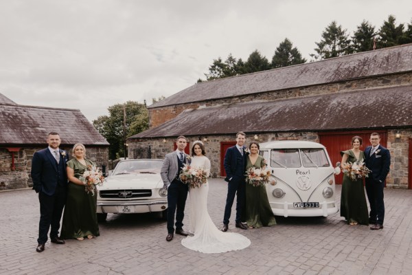 Bride groom bridesmaids and groomsmen stand beside two wedding cars