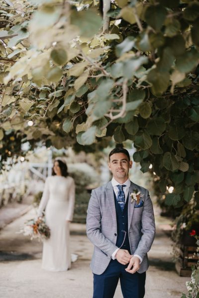 Bride behind groom approaching him in garden