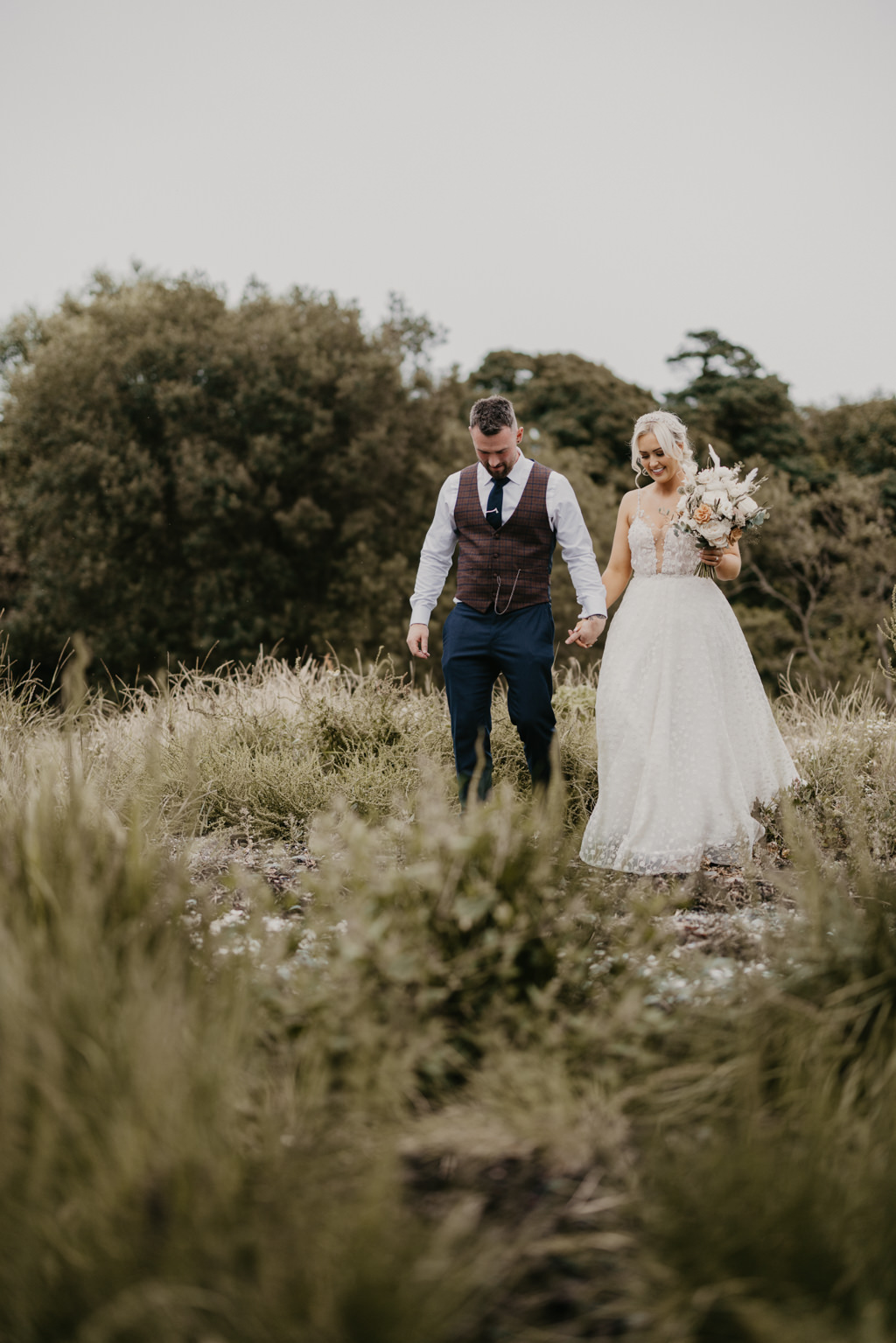 Bride and groom walk hand in hand heading towards beach on grass