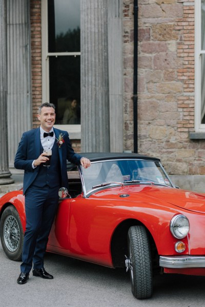 Groom leans on red wedding car holding pint of Guinness