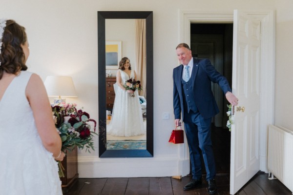 Mirror reflection of bride holding bouquet flowers and father entering room with red gift bag