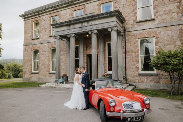 Bride and groom outside venue beside red wedding car