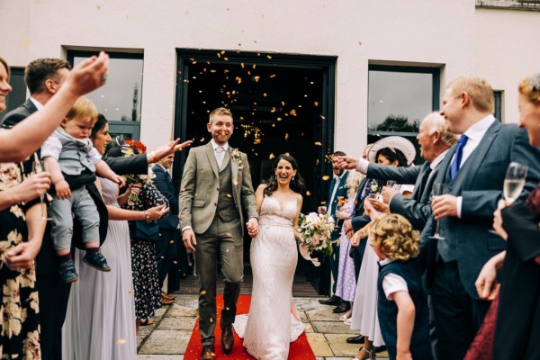 Bride and groom walk the orange carpet surrounded by guests throwing confetti on couple