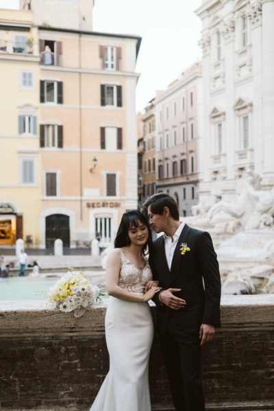 Groom kisses bride on head as they pose in front of fountain