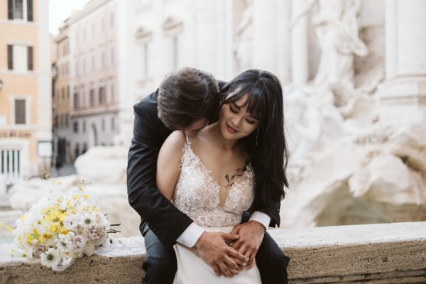 Groom hugs bride from behind they smile in front of fountain