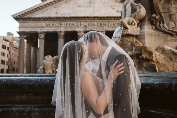 Veil covers both bride and groom as they kiss underneath in front of fountain pillared building