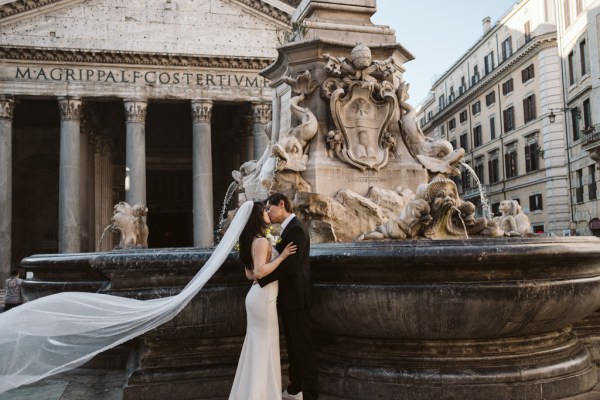 Veil covers both bride and groom as they kiss underneath in front of fountain pillared building