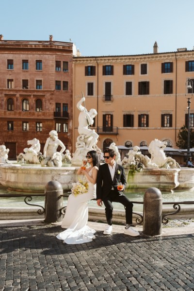 Fountain in background couple bride and groom in foreground drinking Aperol Spritz cocktails sitting down bench