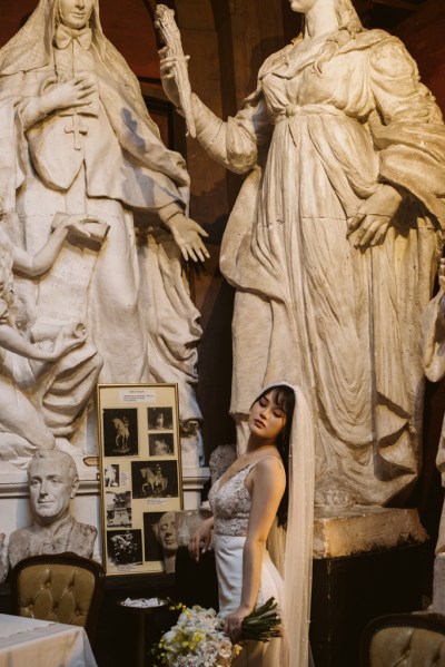 Bride looks above at statues she holds bouquet flowers