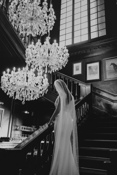 Bride on her own chandelier above standing on staircase portraits in background black and white