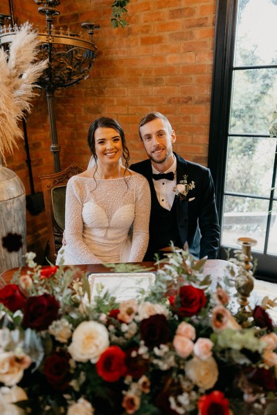 Bride and groom face camera after signing marriage cert on table covered in flowers and roses