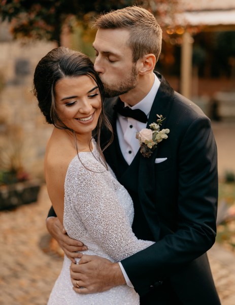 Groom kisses bride on the cheek as she smiles and looks down