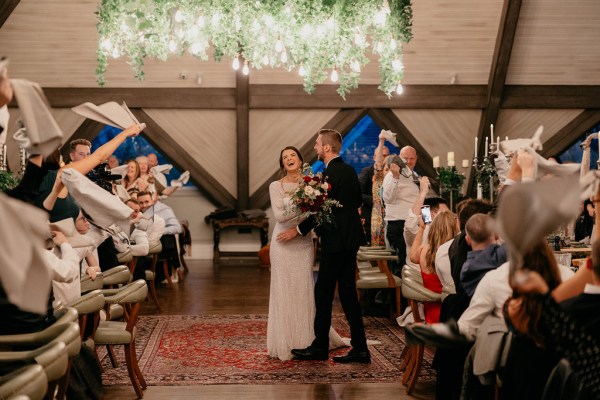 Bride and groom stand in front of guests in dining room/ballroom setting smiling