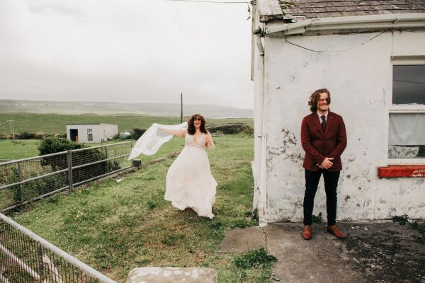 Bride shows off her long dress dancing on grass veil blowing in the wind and groom standing beside wall