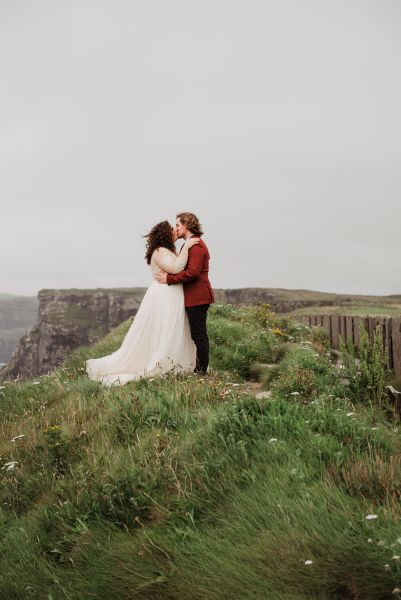 Bride and groom hug embrace on grass cliff edge Irish landscape kissing they kiss