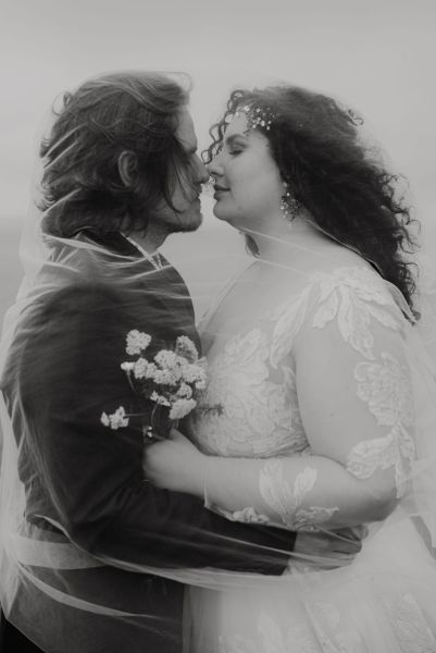 Bride and groom stand on grass setting sea cliff wind is blowing close up black and white