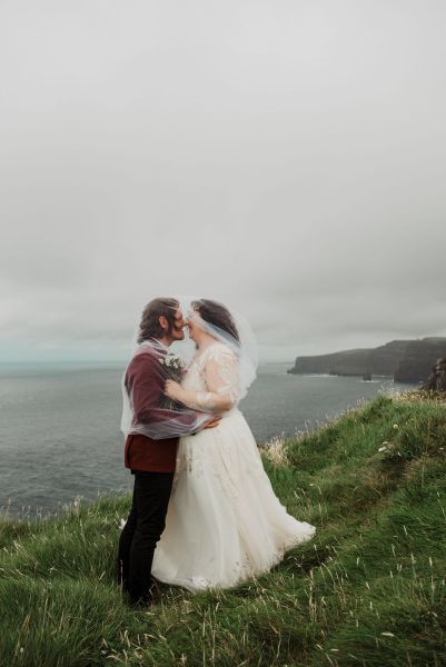 Bride and groom stand on grass setting sea cliff wind is blowing