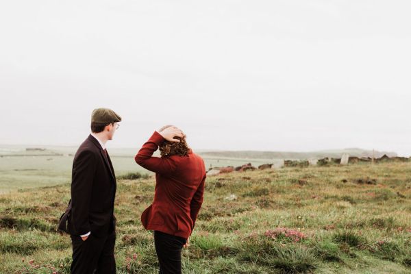 Groom in red suit and groomsman stand on grass cliff setting