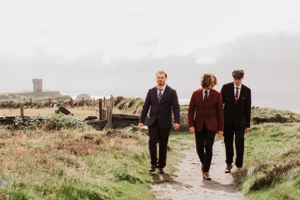 Groom and groomsmen pose laugh on grass cliff setting walking along pathway landscape