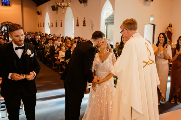 Black and white image of bride and groom at the alter with priest guests in background they are kissing kiss