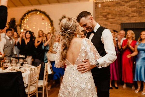 Groom places hand on brides waist in dining room surrounded by guests