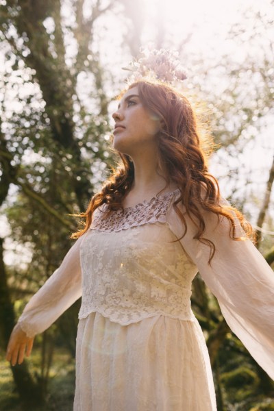 Arms either side of model bride as she wears floral flower headband dances in forest setting