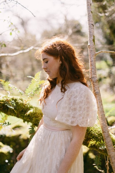 Model bride stands leans against tree bark in forest