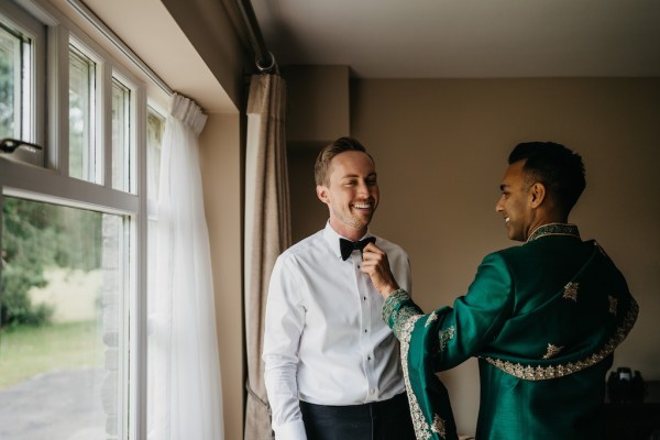 Grooms help each other get ready at window bowtie