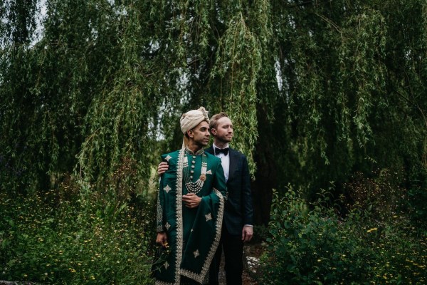 Grooms stand in garden surrounded by trees looking away