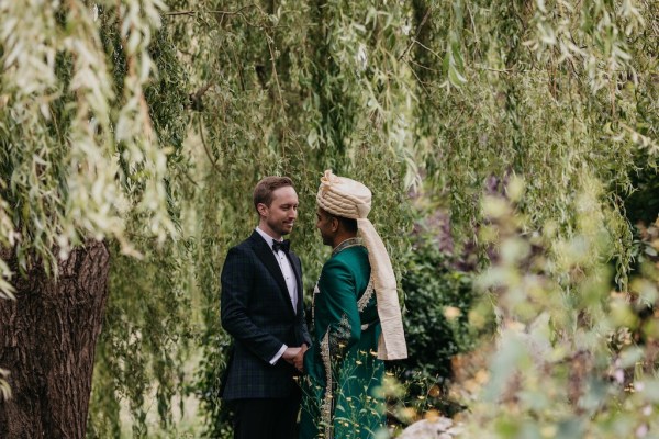 Grooms stand in garden surrounded by trees looking at each other