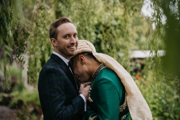 Grooms stand in garden surrounded by trees groom rests head on grooms shoulder