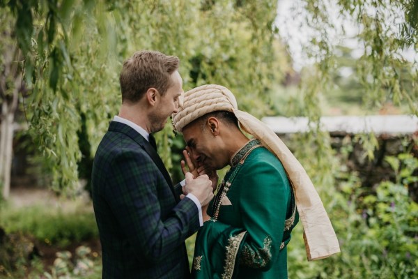 Grooms stand in garden surrounded by trees groom rests head on grooms shoulder