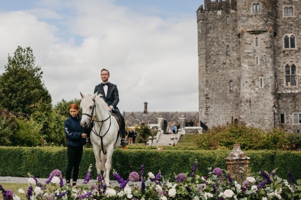 Groom in black suit walking into ceremony on white horse