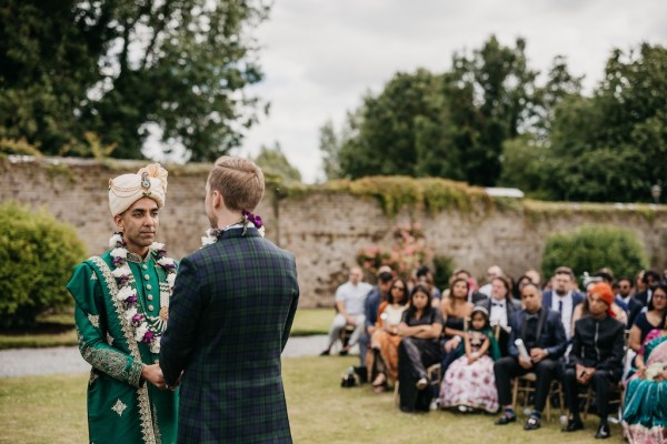 Guests seated during ceremony chairs on grass watching grooms holding hands