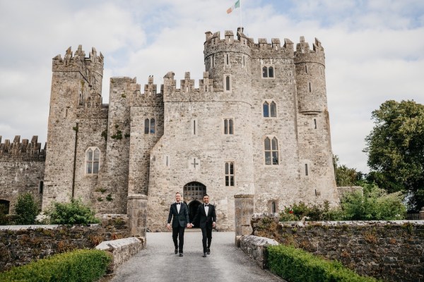 Grooms in black suits walk away from castle on pathway in garden