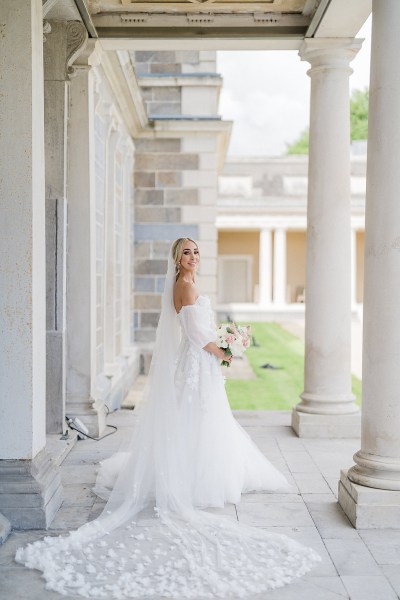 Bride poses beside pillars to wedding venue holding bouquet