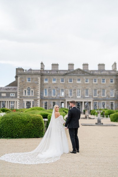 Bride and groom walk along pathway to wedding venue castle