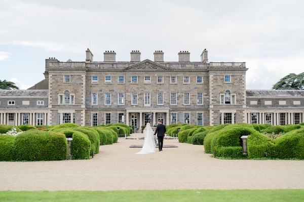 Bride and groom walk along pathway to wedding venue castle